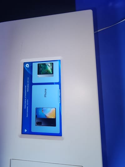The image shows a close-up, angled view of a kiosk screen with a blue interface. The screen displays two product images: a tablet on the top and a smartphone on the bottom. Text in Russian is visible on the left side of the screen, including "Номер устройства" (Device Number) followed by "MT004020424161941", and "Версия программы" (Program Version) followed by "V22.05.18", and "Версия данных" (Data Version) followed by "MT/1.0.2". The text on the right of the bottom phone image reads "Вырезать в избранное" (Cut to favorites). The kiosk is positioned in what appears to be an indoor public space, with a bright blue wall or partition visible to the right of the kiosk. The overall setting is clean and modern. There is no indication of the city of Ushtobe, Kazakhstan in the image.
