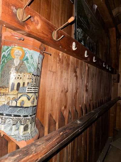 An eye-level, high-angle shot depicts an attic interior. A wooden ladder-like structure is attached to the ceiling, which is also made of wood planks. On the left, partially visible through what appears to be a glass panel, is a poster or sign with text in a foreign language. A wooden beam with several white hooks and a few brass hooks is affixed to the left side of the scene. Below this beam is an elongated ceramic tile or painted object featuring a religious figure with a golden halo, buildings, and the word "MONASTERY" written on its side. The wooden ceiling exhibits varying shades of brown, and the overall lighting is subdued. This appears to be an attic location near Rila Monastery in Rila, Bulgaria.