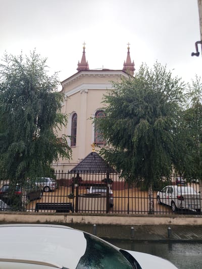 The image depicts a tan building, likely a church or religious institution, with two prominent spires topped with crosses. The building has arched windows and ornate architectural details. In front of the building, a dark metal fence encloses a courtyard area with several parked cars, including a white sedan with a taxi sign on top. Two large trees with dense foliage flank the building, partially obscuring its facade. A small, dark gazebo with a tiled roof is situated within the courtyard, near the fence. The ground appears wet, suggesting recent rain, and the sky is overcast, indicating a cloudy or possibly inclement weather day. A white car is visible in the foreground, its hood and windshield covered in water droplets, further reinforcing the impression of wet conditions. The overall scene suggests an exterior view of a building in a town or city setting. The location context provided is Arad, Romania, and a visible license plate on the white taxi reads "AR 12 MMK". There is also text on the taxi door which is too blurry to read.