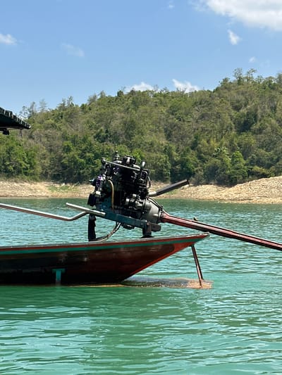 A longtail boat powered by a diesel engine rests on a body of water. The boat is oriented at an angle in the frame with the engine prominently visible. The stern of the boat is a dark reddish-brown with green accents along the edge. A long, metal pole extends into the water.
Behind the boat, a shoreline slopes upwards and is covered with lush, green vegetation and trees. A strip of sandy or gravelly beach separates the water from the foliage. Above the treeline, a blue sky is dotted with a few clouds.
Based on the location in the city of Ban Ta Khun, Thailand, this body of water is likely Cheow Lan Lake.