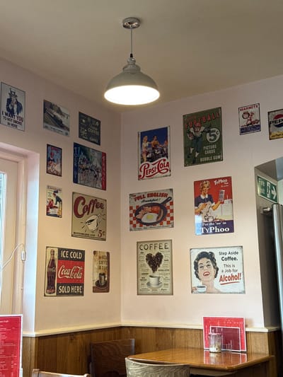 This is an indoor shot of the interior of a café or diner, possibly in Bowness-on-Windermere, United Kingdom. The walls are adorned with vintage-style advertising posters and signs featuring brands like Pepsi, Coca-Cola, Typhoo Tea, Marmite, and Lucozade, interspersed with coffee-themed signs. A light fixture is visible, casting a bright glow. A window is present at the bottom, along with a glimpse of a menu on a table. Some wooden tables and chairs are visible on the right side of the image. There are no people visible in the frame.