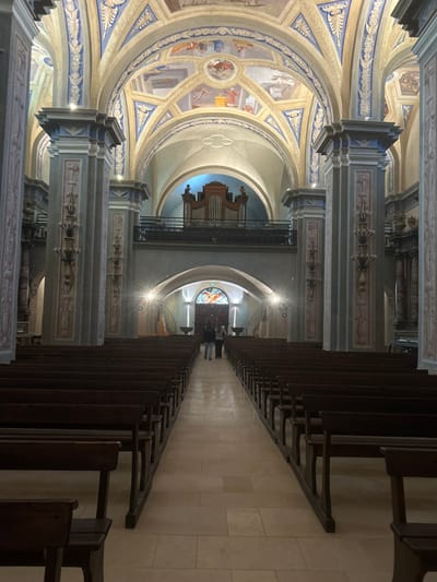 The interior of a church features rows of wooden pews lining a central aisle. High, decorated columns support the arched ceiling with painted frescoes. A large pipe organ is positioned centrally, above a stained-glass window at the far end. The scene is brightly lit, with flares from light sources emphasizing the architecture. Two people stand near the stained-glass window in the distance.