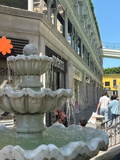 This image captures an outdoor commercial street scene in Gaziosmanpaşa, Turkey, under clear daylight.
In the foreground, a multi-tiered, white marble fountain dominates the lower left, with water actively flowing from its upper basins into the large, greenish-water-filled lower basin. A spherical finial tops the uppermost tier.
The midground features a paved pedestrian area lined with shops. To the left of the fountain, partially visible behind it, a reflection of a person in a white cap appears in a shop window. Further left, an orange, star-shaped logo is partially visible. The shop directly behind the fountain is identified by a prominent black sign displaying "modanisa". A woman in a brown hijab and dark clothing is partially visible near the fountain's left side, looking downward. To the right of the fountain, a young man with dark hair is seen from behind, looking into the fountain's basin. A black metal railing separates the fountain area from the adjacent pedestrian path. On the path, two individuals walk away from the viewer: a man in a white t-shirt and blue jeans, carrying a white shopping bag with an orange logo, is followed by a woman in a light blue button-up shirt and blue jeans. Further along the path, another shop entrance marked "S.O. Chic" is visible.
The background consists of a long, multi-story building running parallel to the street on the left, characterized by numerous classical columns, arched windows, and decorative cornices. Green foliage lines the top edge of this building. A light-colored pedestrian bridge with blue railings spans the upper right portion of the image, connecting to another structure. Beyond the shops, a distinctive yellow building with a dark roof and multiple windows is visible, featuring a black sign that reads "Outlet VAKKO". The sky above is a clear, bright blue.