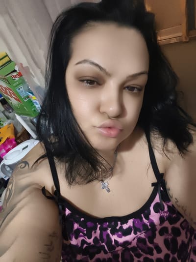 The image shows a close-up, head-and-shoulders shot of a young woman with long, dark hair. She is looking towards the camera with her lips slightly pursed. She is wearing a pink and black leopard-print top with thin spaghetti straps. A silver cross necklace hangs around her neck. She has tattoos on her left arm and shoulder, partially visible. The background is blurred and appears to be an indoor setting, possibly a bedroom, with some items on a shelf. The lighting suggests it might be evening or dimly lit. There is no visible indication of the city of Komotini, Greece in this image.