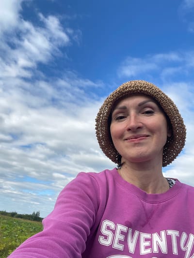 A woman wearing a woven straw hat and a purple sweatshirt with "SEVENTY" printed on it, takes a selfie outdoors. She is smiling at the camera. The background shows a blue sky with white clouds, and a green field with some trees in the distance. The location is the city of Dresvyanovo, Russia.