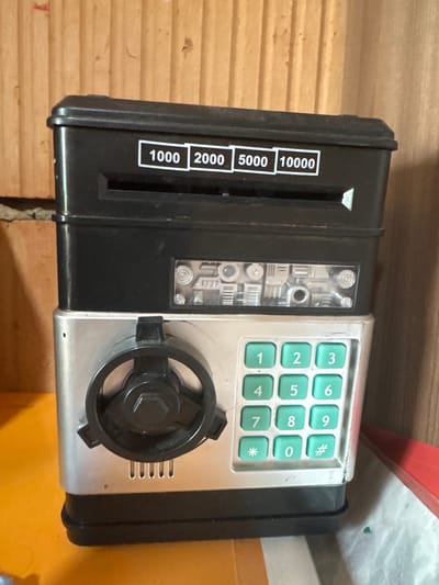 A black and silver toy ATM is placed on an orange surface in front of a wooden-paneled wall. The ATM has a coin slot at the top and a numerical keypad with green buttons. There is a dial on the front of the ATM. The background is a wooden wall with vertical planks. The image is a medium shot, taken from a slightly elevated angle.