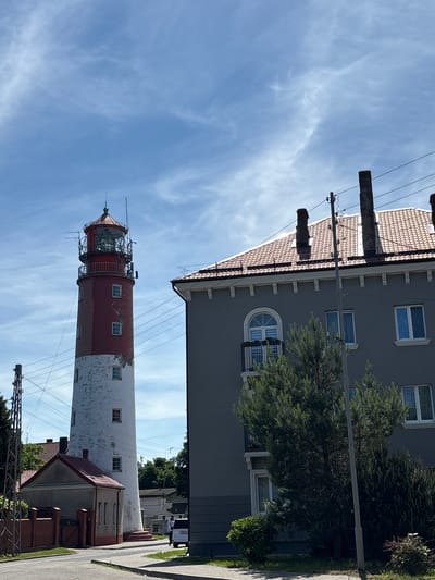 The image shows a lighthouse in Baltiysk, Russia, a port city located on the Baltic Sea. The lighthouse is a tall, cylindrical structure, painted white and red, with a lantern room at the top. It stands next to a modern building with a red tiled roof and several windows. A smaller, gabled building with a red roof is situated at the base of the lighthouse. The scene is set against a backdrop of a blue sky with scattered white clouds. Power lines and a street light are visible in the foreground, and a white van is parked on the side of the road. The vegetation includes trees and bushes. The overall impression is one of a coastal town with a mix of historic and contemporary architecture.