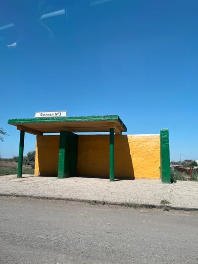 A bus stop shelter with a yellow and green color scheme stands under a clear blue sky. The shelter has a flat, green-painted roof supported by green poles. The back wall of the shelter is painted yellow and textured. A sign above the entrance reads "Филиал №3" in black lettering on a white background. The ground in front of the shelter is paved with gravel, and a road is visible in the foreground. To the right, a green pillar stands against the yellow wall. In the background, there are some bushes and houses visible. The location is identified as Il'inka, Russia.