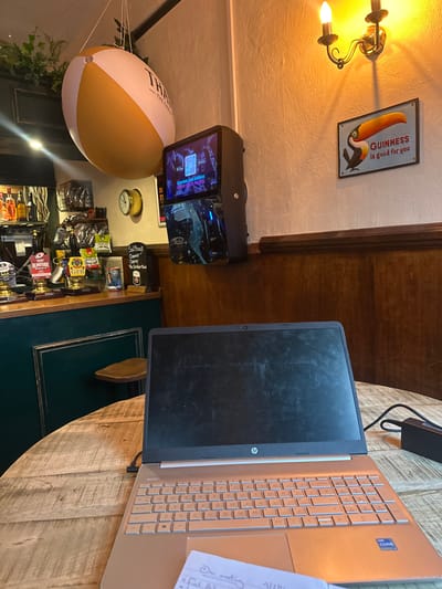 This image depicts the interior of a pub identified as "Salisbury's Favourite Classic 10s Corner Bar" in Salisbury, United Kingdom.
In the foreground, an open HP laptop with a gold-colored chassis and a dark, reflective screen is positioned on a round, light-colored wooden table with a visibly textured grain. A black laptop charger block rests to the right of the laptop. Partially visible beneath the laptop's touchpad is a small white slip of paper with handwritten text, including "Our meeting" and "2/24".
The midground features a dark green wooden bar counter with a gold trim along its base on the left. Several beer taps are visible on the bar top, including labels "BLINDSIDE" and "GFB". Behind the bar, bottles of spirits are arranged on shelves, and bags of snacks are suspended in a basket. A single wooden bar stool is partially visible in front of the counter. Above the bar, a large, inflatable beach ball, white and tan, hangs from the ceiling. A portion of text, "THAT", is visible on the beach ball, suggesting "THAT family-friendly pub".
To the right of the bar area, a black wall-mounted digital display unit is fixed to the wall. Its upper screen shows a QR code and the text "CONTROL THE JUKEBOX FROM YOUR PHONE". The lower screen displays a dynamic blue graphic, possibly an animation or advertisement. The wall behind these objects is painted white above a dark wooden paneling. A brass-colored wall sconce with two candle-shaped bulbs emits warm light. Below the sconce, a rectangular sign features a toucan and the slogan "GUINNESS is good for you". No individuals are visible in the scene.