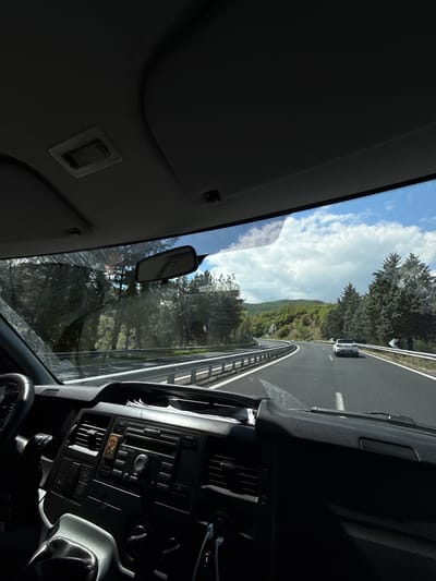 The image presents a view from inside a vehicle, looking out through the windshield onto a scenic road and landscape. The vehicle is on an asphalt road with white lane markings and a metal guardrail visible on the right. Ahead, another white vehicle, possibly a van, can be seen. The road appears to wind through a mountainous or hilly region, flanked by dense green trees, including conifers, and lush vegetation under a bright blue sky with scattered white clouds.

Inside the vehicle, the dashboard is visible on the right, featuring a car radio/control panel with text such as "AM/FM", "CLOCK", "TA", "VOL", "CD", "AUX", and "FORD 6000 CD", indicating a Ford vehicle. A small, colorful religious icon is affixed to the dashboard below the radio. Parts of the steering wheel are visible at the bottom right. Sun visors are seen above on the left, and a rearview mirror hangs in the upper center of the windshield, which has some visible dust or marks.

The scene depicts a clear, sunny daytime with bright light entering the vehicle. The overall impression is that of a road trip or journey through a natural, possibly mountainous, landscape, aligning with the Strymoniko Municipal Unit, Greece, context.