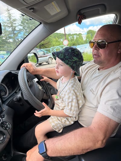 A man wearing sunglasses and a beige t-shirt sits in the driver's seat of a car, with a young child on his lap. The man's left hand rests on the steering wheel, and the child's hands are also on the wheel. The child wears a patterned bucket hat and a checkered shirt. The car's interior is visible, including the dashboard and steering wheel. Outside the car, trees and a blue sky with clouds can be seen. A decorative ribbon with orange and black stripes is tied to a handle above the windshield. The scene is likely captured in the city of Kul'dur, Russia.