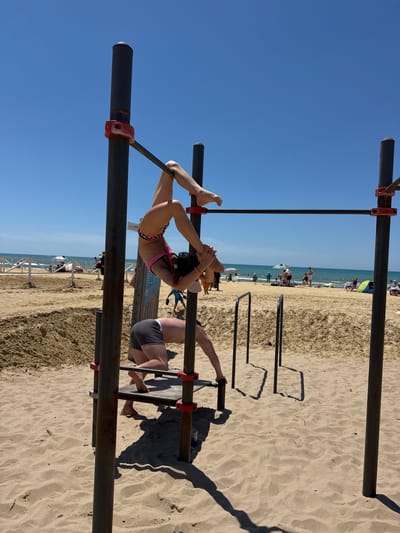 The image depicts an outdoor exercise area on a sandy beach, likely located in Natukhayevskaya, Russia, with the Black Sea visible in the background under a clear blue sky. Two individuals are engaged in physical activity on a metal workout structure. One person, wearing a pink and leopard-print bikini, is suspended upside down, holding onto a horizontal bar with their hands while their legs are bent. Another person, in grey shorts, is positioned below, leaning forward and grasping a different part of the structure. The beach is populated with other beachgoers, umbrellas, and tents, indicating a typical summer day. The workout equipment consists of vertical poles and horizontal bars, set up on the sand.