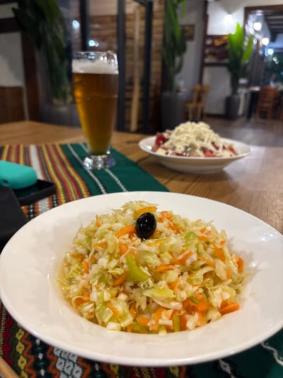 A close-up, eye-level shot shows a white bowl filled with coleslaw topped with a single black olive. The coleslaw consists of shredded cabbage, shredded carrots, and chopped green peppers in a light dressing. In the background, a tall glass of beer with a foamy head sits on a table. Further in the background, another white bowl containing a salad topped with shredded white cheese is visible. The scene appears to be set in a restaurant in Smilyan, Bulgaria, with out-of-focus plants and furniture in the background. The table is covered with a green placemat with a colorful, geometric pattern.