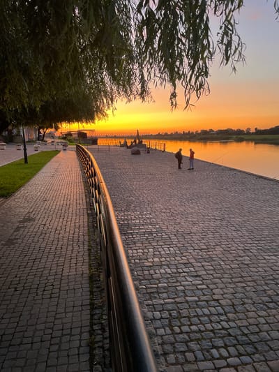 The image depicts a cobblestone promenade along a wide river at sunset. The sky is ablaze with vibrant orange and yellow hues, reflecting on the calm water. Lush green trees, likely weeping willows with their characteristic drooping branches, partially frame the scene from the top left. Two individuals are visible on the promenade, one in a dark jacket and the other in a bright orange jacket, both appearing to be fishing with rods. Several fishing rods are strategically placed along the riverbank. In the background, a bridge spans the river, and the opposite bank is lined with trees. The promenade is a mixture of paved walkways and cobblestone surfaces, with decorative lampposts and planters along the edge. The scene is set in Sovetsk, Russia, specifically on the embankment of the river.