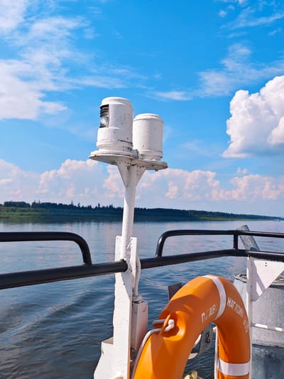 The image shows a view from the deck of a boat on a body of water, likely the Podkamennaya Tunguska River in Russia, on a sunny day. The sky is blue with scattered white clouds. In the foreground, there is a white railing and a white pole with two white cylindrical objects on top, possibly navigation lights. To the right, an orange life preserver is attached to a railing. In the background, a lush green forested shoreline is visible across the water. The water is calm with ripples. There are no people visible in the image.