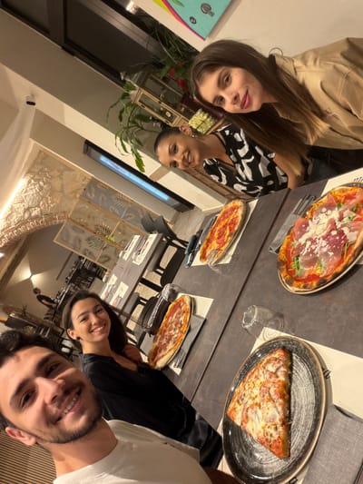 This indoor photograph captures four individuals, two women and two men, dining at a restaurant or pizzeria in Corsico, Italy. The image is taken from a slightly elevated, upward-looking perspective, capturing the faces of the patrons and the table spread.

On the left, two women are seated. The woman closer to the camera has long dark hair and wears a light-colored jacket, looking directly at the viewer with a slight smile. Beside her, another woman with dark hair and a black and white patterned top smiles towards the camera.

The table in the foreground holds several pizzas. A large pizza with prosciutto, arugula, and shaved cheese on an oval plate is prominent towards the top-center. To its right, a single large slice of a classic cheese pizza rests on a dark, textured plate. Another smaller, round pizza with red sauce and cheese is visible further down the table.

On the right side of the image, a woman with dark hair and a black top smiles, positioned behind a man with a beard and a white t-shirt, who is also smiling directly at the camera. Their expressions convey a pleasant, social gathering.

The setting features dark tables and chairs. The background includes decorative screens with leaf patterns, various green potted plants, and sections of exposed brick or stone walls, suggesting a modern yet rustic interior design. Warm, artificial lighting illuminates the scene, indicating an evening meal. Faint, illegible text is visible on some of the paper table settings.