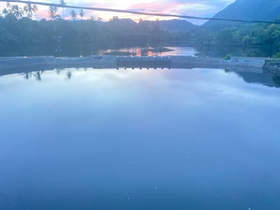 The image shows a calm body of water, likely a reservoir or pond, with a concrete structure running across it. The structure has several openings or gates. In the background, there are lush green trees and hills, with palm trees visible on the left. The sky is a gradient of blue and pink, suggesting either sunrise or sunset. There are no people or animals visible in the image. The overall scene is tranquil and natural. The location of Aneuk Paya, Indonesia, is provided contextually but is not directly depicted within the visual elements of the image.