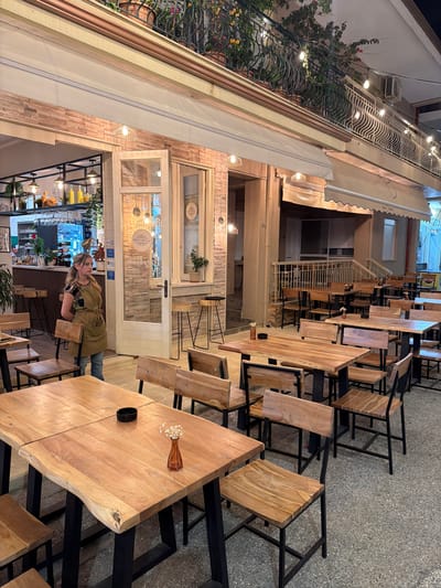An outdoor seating area of a restaurant in Asprovalta, Greece is depicted at night. A woman wearing an apron stands near an open doorway, facing the viewer with a neutral expression. The restaurant features wooden tables and chairs with black metal frames. Several tables are set with small vases of flowers and ashtrays. String lights are visible hanging from the building and balcony above, illuminating the scene. The background includes the interior of the restaurant, a bar area, and additional seating. The overall ambiance suggests a quiet evening at a casual dining establishment.
