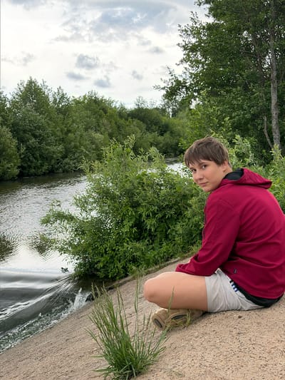 A photograph taken outdoors depicts a young person crouching at the edge of a concrete structure beside a body of water. The young person, facing the camera, is wearing a maroon hooded jacket and light-colored shorts with tan shoes. They are positioned on the right side of the frame. The concrete structure serves as a boundary, likely for the water. To the left, the water is surrounded by dense green vegetation, appearing to be trees and bushes. The sky is visible at the top left, showing overcast conditions with clouds. The image is oriented so that the ground appears on the right and the sky on the left. The location is Nizhnii Vishur, Russia.