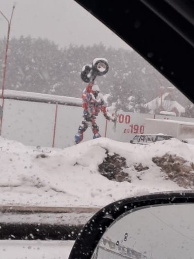 This image depicts a snowy, overcast daytime scene in Staraya Kupavna, Russia, viewed from inside a vehicle. The central subject is a large, humanoid statue, predominantly colored red, white, and blue, constructed from tires and other components, resembling a robot or a Transformer. This statue stands atop a significant mound of snow and holds two black tires linked together above its head with its right arm. Its left arm is bent and held forward.
In the mid-ground, behind the statue, a low, white structure, possibly a wall or fence, is visible, marked with partially obscured red characters "00" and "190." A dark vehicle is partially visible behind this structure, to the right of the statue, mostly covered in snow. The background features a dense line of snow-covered evergreen trees beneath a grey, snow-falling sky. The ground throughout the visible area is covered in substantial snow. In the foreground, the black frame of a vehicle's side mirror, speckled with moisture or snow, occupies the lower right corner. Snow is actively falling across the entire scene.