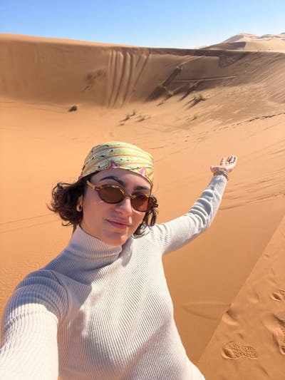 A single female individual is centrally positioned in the foreground, captured in what appears to be a selfie in a desert setting near Rissani, Morocco. She has dark, curly hair, partially covered by a light green and pink patterned headscarf. She wears tortoise-shell sunglasses, small gold-colored hoop earrings, multiple thin gold-colored bracelets on her left wrist, and a light-colored, ribbed turtleneck sweater. Her left arm is raised and extended to her side, with her palm open in a gesturing motion, and she displays a slight smile. The background is dominated by extensive orange sand dunes exhibiting various textures, shadows from direct sunlight, and scattered sparse vegetation. Footprints are visible in the sand to the lower right. The sky overhead is clear and blue, indicating daytime. The scene's characteristic large orange dunes are consistent with the Erg Chebbi desert landscape.