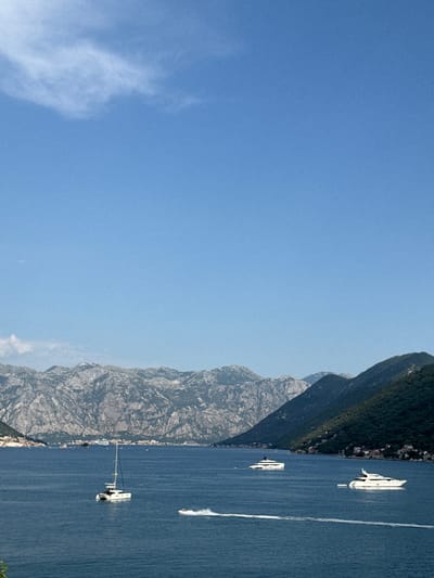 This is a vertically oriented, eye-level, medium shot of a serene lake with steep, tree-covered mountains on one side and a clear blue sky on the other. The lake is a deep blue, reflecting the sky, and a few white boats are visible on its surface. One boat, closer to the viewer, appears to be a white yacht with several people on deck. Further in the distance, another white boat, possibly a speedboat, is creating a white wake as it moves across the water. Along the base of the mountains, a small, picturesque village can be seen nestled between the water and the steep slopes. The sun is shining brightly, casting a soft light on the scene, suggesting it is a clear, warm day. The overall impression is one of tranquility and natural beauty. The image was taken from a boat or a vantage point on the water, looking towards the shore and the mountains.