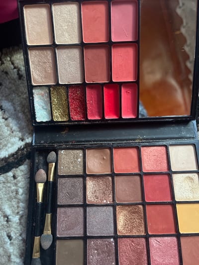 This is a close-up, overhead shot of an open makeup palette. The palette is black and divided into rectangular sections, each containing a different shade of eyeshadow or blush. The left side of the palette features a mirror at the top, reflecting a blurry, warm-toned image. Below the mirror are several rows of makeup pans, including shades of pink, coral, and red, with some glittery textures. The right side of the palette is filled with eyeshadow pans in various shades of brown, rose gold, and shimmer.
At the bottom of the right side of the palette, two makeup applicators with gold handles and grey-brown sponge tips are visible. The palette is resting on a textured surface that appears to be a rug or carpet with a black and white patterned design. The lighting in the image is warm, suggesting an indoor setting. There is no visible text in the image. The overall impression is of a well-used makeup product. The location context of Ташлак, Kyrgyzstan, is not visually discernible from the image itself.
