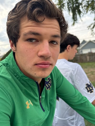 An outdoor, eye-level image depicts two young adult males in South Bend, United States. The primary subject, in the foreground and positioned left of center, is a male with brown wavy hair and a neutral expression, looking directly at the camera. He wears a green quarter-zip athletic top with a gold Under Armour logo on the left chest and a navy and gold "ND" (University of Notre Dame) logo on the right chest. Partially obscured behind and to the right of the primary subject is a second male, seen from the back, wearing a white long-sleeved athletic shirt featuring a dark "ND" logo on the upper left sleeve. The background is out of focus, showing residential-style buildings with light-colored siding and dark roofs, a section of fencing, and green foliage under an overcast sky.