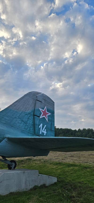 This image shows the tail section of a vintage airplane, likely a Soviet-era military aircraft, situated outdoors in a grassy field. The plane's tail is painted a teal-blue and features a red five-pointed star insignia with white trim. Below the star, the number "14" is prominently displayed in white. A portion of the aircraft's wing is visible in the foreground, resting on a concrete support. The background consists of a dense line of trees and a sky filled with dramatic, partly cloudy formations. The setting appears to be a historical monument or museum, given the prominent display of the aircraft. The location is identified as the Museum of the Carpathian-Slovak Uprising in Vyšný Komárnik, Slovakia. No people are present in the image.