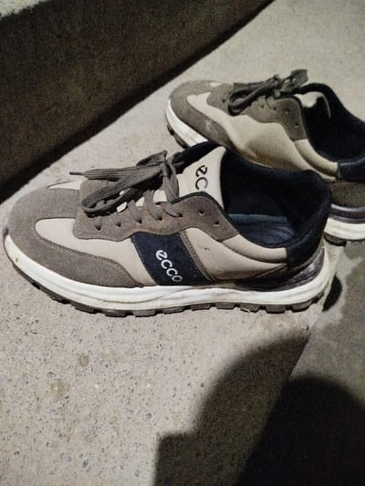 The image displays two pairs of Ecco brand sneakers resting on a grey, textured concrete surface in Yakkabog, Uzbekistan. The shoe in the foreground is in sharp focus, oriented diagonally across the lower left and center. It features a design incorporating light beige and darker brown/grey panels, with a prominent dark blue or black stripe along the side. The word "ecco" is clearly visible in white text on this dark stripe, positioned vertically, and also horizontally on the inside collar of the shoe. The sneakers have white laces and a white midsole with a dark, ridged outsole.
The second shoe is partially visible in the upper right, slightly out of focus, and appears to be of the same model and color scheme. Distinct shadows are cast across the concrete surface, particularly a large dark shadow in the upper left, suggesting low light conditions such as dusk, dawn, or artificial illumination. No people are present, and no specific activity is taking place; the shoes are simply depicted at rest. The concrete surface appears dry and shows minor imperfections consistent with an outdoor setting.
