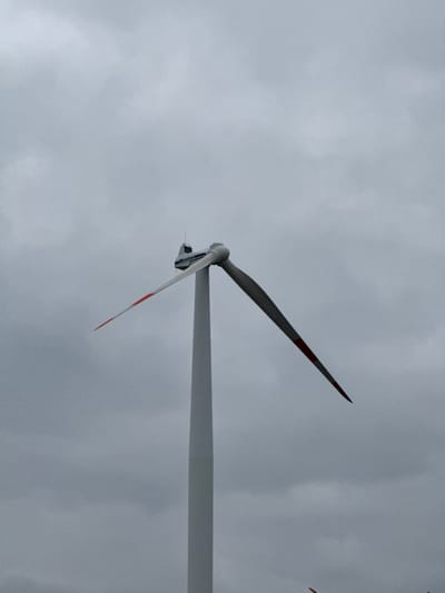 This image depicts a Vestas wind turbine located in Dornstadt, Germany, positioned against an overcast, uniform gray sky. The turbine consists of a white, tapering cylindrical tower supporting a white nacelle. The "Vestas" logo in blue text is visible on the side of the nacelle. Two rotor blades are visible; one extends upward and to the left from the nacelle, featuring a red tip, while the other extends downward and to the right, also with a red tip. The blades appear stationary. No people, other structures, or ground features are present in the frame.