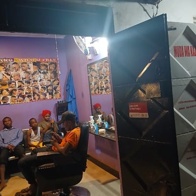 The image depicts the interior of a barbershop located in Mji Mpya, Tanzania, viewed through its partially open entrance at night, indicated by artificial lighting.
Inside, five individuals are present. In the foreground, a male in an orange t-shirt and dark cap is seated in a black hydraulic barber chair, facing a large mirror, and is operating a smartphone. To the left, on a waiting bench, sit three individuals: an adult male in a light blue shirt, a male child in a white tank top, and a female in a dark green top with a red headwrap who is also using a smartphone. A fifth individual, matching the description of the female with the red headwrap, is reflected in the mirror, seated behind a counter containing various grooming products.
The barbershop's walls are light purple. A large poster displaying men's hairstyles, labeled "ING - WILD & FREE," is affixed to the left wall. A blue towel hangs nearby, and a white wall-mounted fan is visible. The entrance features a dark, possibly metal, door with a diamond pattern. A red and white sign on the door's interior states "Lipa kwa Simu hapa" (Pay with phone here), with mobile payment logos. Beyond the door, an exterior wall with geometric patterns displays red text, including "MUDA WA KAZI" (Working Hours). The interior is brightly illuminated by overhead artificial light sources.