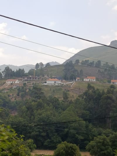 The image displays a hillside landscape in Terras de Bouro, Portugal. Several houses with orange-tiled roofs are scattered across the incline, interspersed with patches of dense green foliage and trees. A construction crane is visible on the upper part of the hill, suggesting ongoing building activity. Power lines stretch across the frame, dividing the sky from the landscape. The overall scene is one of rural development on a hilly terrain under a cloudy sky.