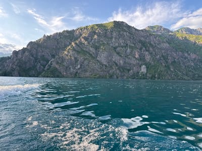 The image depicts a serene natural landscape in Arkit, Kyrgyzstan, likely captured during the daytime. The foreground is dominated by the choppy, dark blue water of a lake, with white foam and ripples indicating movement, possibly from a boat. The sun glints off the water's surface, creating bright highlights.
In the background, a majestic, rugged mountain range rises steeply from the water's edge. The mountains are characterized by their rocky, textured surfaces, with numerous sharp peaks and crevices. Patches of green vegetation, including trees and shrubs, cling to the slopes, especially in the higher, more verdant areas. The summit of the largest mountain is covered in lush green grass and sparse trees.
The sky above is a clear, bright blue, punctuated by wispy white clouds that are illuminated by the sun. The light suggests it is either late morning or late afternoon, casting a warm glow on the scene. The overall mood is peaceful and awe-inspiring, highlighting the natural beauty of the mountainous terrain and the tranquil lake. There is no visible text in the image.
