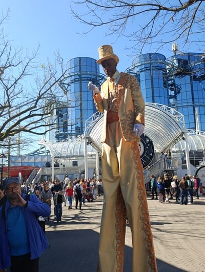 The image captures a scene in Rust, Germany, in front of the silver-colored Geisterschloss attraction with the blue mirrored Euro-Tower building in the background. The area is populated by a crowd of people, many in casual clothing. In the foreground is a man on stilts, dressed in a gold-colored, patterned suit and top hat. To the left is another man in a blue jacket and cap, partially obscured and looking towards the stilt walker. The environment is an open, paved area with bare trees framing the scene. The overall impression is of a public space in a tourist destination on a sunny day.