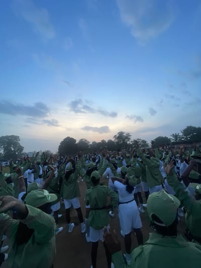 An outdoor, ground-level, full shot captures a large group of young adults, presumably participants in a National Youth Service Corps (NYSC) program, engaged in physical activity under a pale blue, cloudy sky at dawn. The individuals, dressed in matching green shirts, white shorts, and green caps, are spread across a dirt field bordered by trees and a distant structure with a fence. Their arms are raised in unison, suggesting a coordinated exercise or dance. The scene is one of communal activity and discipline, characteristic of a structured training environment. The low light suggests early morning.