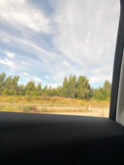 The image is an outdoor scene, viewed through what appears to be a car window. In the foreground, a dark, textured surface, likely the interior of a car door or armrest, occupies the bottom third of the frame. The middle ground shows a blurred landscape with trees and vegetation, suggesting a natural or semi-natural environment. Above the treeline, a blue sky with wispy white clouds is visible. The overall impression is of a journey through a rural or suburban area. The location is identified as Skumparp, Sweden. There are no people or specific actions depicted in the image.