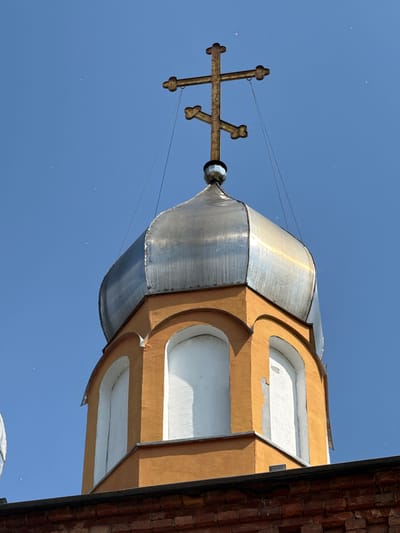 The image shows the dome and cross of a church in Chudovo, Russia, against a clear blue sky. The dome is metallic and appears to be made of silver-colored material. The cross is made of rusted metal and is positioned at the apex of the dome. The lower part of the structure is painted orange and features arched windows with white interiors. The bottom edge of the image shows a section of a red brick building. There are no people visible in the image.