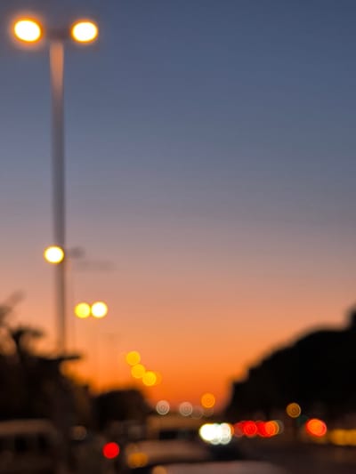 The image depicts a blurred, bokeh-heavy scene of streetlights and vehicle lights against a gradient sky. The sky transitions from a deep blue at the top to a warm orange and pink hue towards the horizon, indicating a sunset or dusk.

The most prominent subjects are several streetlights, appearing as bright, out-of-focus orbs of yellow light. One streetlight in the upper left has two distinct lights, while another lower down has a single, bright light. Further into the scene, a series of smaller, colorful orbs represent vehicle lights, including red taillights and white headlights, suggesting a road or highway at nightfall. Dark, silhouetted shapes on the right side of the image could be trees or buildings.

The overall impression is one of a city or suburban environment at twilight. The blurry nature of the image creates a dreamlike or atmospheric quality, emphasizing the lights and the sky. There is no visible text in the image. The focus is on the abstract beauty of artificial lights against the natural backdrop of a colorful sky at the end of the day. The scene evokes a sense of calm and perhaps the transition from day to night, common in urban settings.