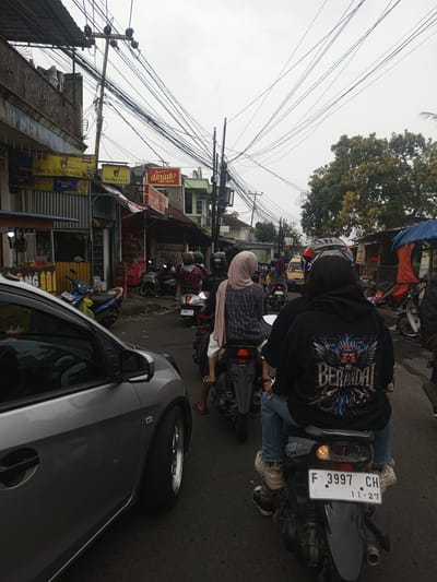The scene is depicted in the city of Houaythat, Laos, on an overcast day, capturing a busy urban street with vehicular traffic. In the immediate foreground on the left, the front-left portion of a silver compact hatchback, featuring custom five-spoke wheels and a dark tire, is visible. To the right of the car, a black scooter is positioned, occupied by a rider viewed from behind. This rider wears a black hooded sweatshirt with a multi-colored graphic depicting a skull and wings above the text 'THE BERANDAL', blue jeans with distressed cuffs, and light-colored sneakers. The scooter's rear displays a license plate, F 3997 CH, with an expiration date of 11.27. To the immediate left of this scooter, another rider, wearing a light pink hijab and a blue plaid shirt, is also on a scooter.
Further down the street, multiple motorbikes and scooters, each with riders typically wearing helmets, are observed in a queue. A white scooter with license plate F 4936 WQC is visible. In the background, a yellow van is discernable.
Along the left side of the street, a row of commercial buildings features various storefronts and prominent signage, including multiple yellow banners for 'SIDOMUNCUL', a red sign for 'akriuk FRIED CHICKEN', and a 'JAMU SELOGAN' banner. Some buildings exhibit corrugated metal cladding and a weathered, ornate metal railing on an upper level. A blue scooter is parked on the sidewalk near these establishments. A tall wooden utility pole stands prominently on the left, burdened by a dense network of black overhead electrical and communication wires that span across the street and into the sky. On the right side of the street, behind the moving traffic, additional commercial structures are present, along with a stall covered by blue and orange tarps. A large tree with full green foliage is situated on the right, further in the background. The road surface is asphalt.