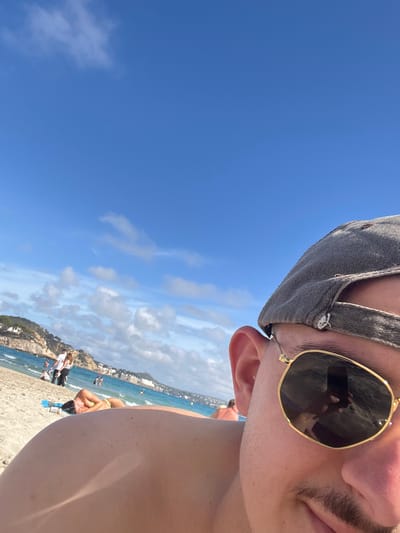 A selfie-style photograph presents a close-up view of a person wearing a gray cap and gold-rimmed sunglasses, taken against a backdrop of Peguera, Spain. The foreground is dominated by the person's face, partially visible, with focus on their ear and part of their cheek and upper lip. The sunglasses reflect the beach scene. The background reveals a beach with several people sunbathing, walking, and swimming in the sea. A building is visible on a cliffside that extends into the water. The sky above is clear blue with scattered white clouds. The overall composition is tilted, providing a dynamic and unique perspective of the beach setting.