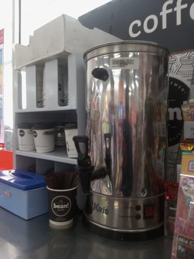 The scene depicts a beverage station on a reflective counter surface, likely within a commercial establishment in Naung Bo Oe, Myanmar. A large, cylindrical Kris-branded stainless steel hot water or beverage dispenser occupies the central and right foreground, featuring two spigots (one prominent on the left) and a base control panel with "Heating" and "Keep Warm" indicators and a red power switch. To its left, a white multi-tiered shelving unit holds multiple white disposable cups with black lids. Directly in front of the dispenser, a black paper cup with the "bean! spot" logo is filled with numerous brown stir sticks. A light blue rectangular plastic container with a lid rests on the counter to the left of the stir stick holder. The background reveals a partial black sign with white text, showing "coffe", and shelves stocked with various packaged retail products, including items like "Creamy Tea" and "Mie Goreng". No individuals are present in the image.