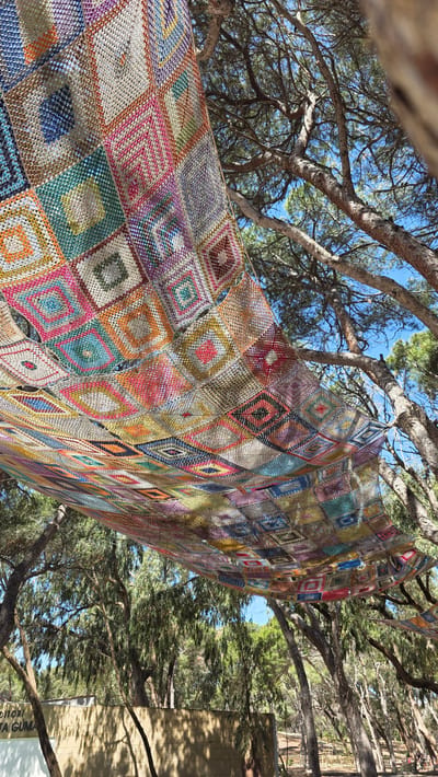 A low-angle, full shot shows a large, crocheted blanket hanging between tree branches under a sunny, blue sky. The blanket is made of numerous square, diamond patterned crocheted panels of various colors, including red, pink, yellow, green, blue, and brown. The blanket appears to be draped or suspended in a natural environment. The tree branches are mostly bare, though with some green foliage visible. In the background to the right, there is a beige building with the inscription "Auditori". The image was taken in Guardamar del Segura, Spain.
