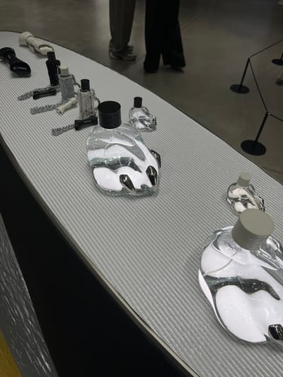 The image depicts an indoor display at a venue in 성동구, South Korea. A long, light-grey display platform with a ribbed, textured surface is angled diagonally across the frame. Arranged on this platform are numerous clear glass bottles of varying sizes, characterized by irregular, organic, or melted shapes with internal white and dark grey/black abstract forms, some resembling claws. Several bottles are topped with black caps, while others have grey caps. Also present are multiple smaller, cylindrical objects with attached silver-colored chains, in black, white, and speckled grey/white patterns, positioned in a line on the platform. Three larger, irregularly shaped objects, two black and one white, resembling stylized footwear or sculptural elements, are situated on the far left of the display. In the background, two individuals are partially visible from the knees down, standing on a dark, polished floor. One person wears light-colored trousers and light-colored shoes, while the other wears dark, wide-leg trousers. Black stanchions with circular bases are visible further back on the dark floor, suggesting a barrier or designated viewing area.