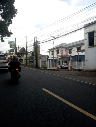 This image captures a daytime street scene, reportedly in the city of Ura-Dozhi, Bhutan, under an overcast sky. The foreground is dominated by a dark asphalt road with a solid yellow line visible on its right side. A person wearing a yellow helmet and dark jacket is riding a dark-colored scooter or motorcycle away from the viewer on the left side of the road, its red taillight illuminated. To the left of the scooter, the rear section of a white SUV is partially visible.
On the right side of the road, a white, two-story building with a gray roof and brown-framed windows stands behind a white fence with red brick pillars. A distinctive red metal archway or decorative element is visible above its entrance. A white van is parked within the fenced area in front of this building. A street light with a solar panel is also present on the right side.
The left side of the road features a green tree with dense foliage. Numerous utility poles support a dense network of overhead wires that crisscross the upper frame. Several commercial signs are visible, including a prominent green sign and a tall, multi-panel vertical sign displaying various colorful graphics and text. Behind these signs, a building with a traditional dark, peaked, tiled roof and wooden architectural elements is discernible.