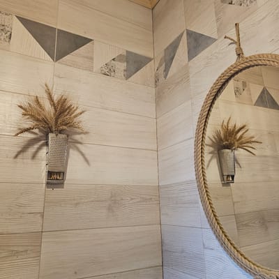The image shows a close-up view of a tiled floor with a wood-like pattern. A circular mirror with a thick rope frame is partially visible at the top of the image, hanging above the floor. In the reflection of the mirror, a small metal grater is seen filled with dried wheat stalks. On the floor, directly below the mirror's reflection, an identical arrangement of a metal grater filled with dried wheat stalks is placed. The floor tiles feature a geometric pattern of triangles in various shades of beige and grey, some with a subtle script-like texture. The lighting suggests an indoor setting, possibly during the day, casting shadows from the wheat stalks and grater onto the floor. The overall impression is one of rustic, decorative arrangement. No people are visible in the image. The location is identified as Vybolovo, Russia.