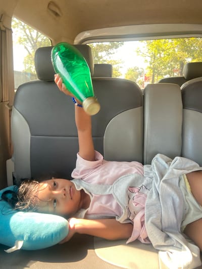 A young girl lies on a car seat with a blue plush toy under her head. She is wearing a pink shirt and grey shorts. Her right arm is raised, holding a green plastic bottle with a blue beaded bracelet on her wrist. The car is likely in motion, with trees and sunlight visible through the windows. The interior of the car appears to be made of grey and beige upholstery.