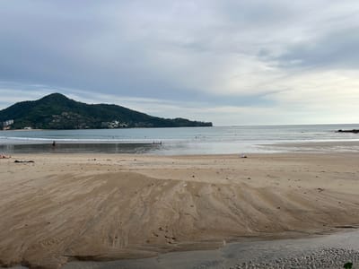 A wide shot of a beach in Ban Bang Thao, Thailand, captures a serene, overcast day. The foreground is dominated by wet sand, showing intricate patterns from receding water. In the mid-ground, the calm sea gently laps the shore, and a few individuals are visible in the shallow water, some standing and others appearing to be playing. Further back, a verdant, tree-covered hill slopes down to the water's edge, dotted with buildings. The sky above is a canvas of muted grey clouds, with a hint of brightness on the horizon suggesting a clearing. The overall impression is one of tranquility and natural beauty.