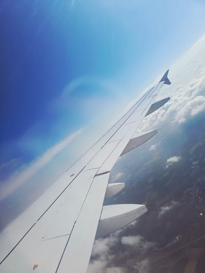 An aerial photograph captures a sweeping view from an airplane window, prominently featuring the aircraft's white wing extending diagonally across the frame. The wing, equipped with a distinct winglet at its tip and multiple engine nacelles along its leading edge, shows intricate panel lines, control surfaces, and several small, yellow/gold-colored attachments. Faint alphanumeric markings, such as "D121" on an engine nacelle, are barely discernible, along with other indiscernible technical text on the wing's surface.
Below the wing, a patchwork of green and brown agricultural fields, interspersed with scattered urban developments, is visible through breaks in a layer of white cumulus clouds. The bright blue sky in the upper left, marked by a gentle sun glare, indicates daytime conditions. The horizon appears significantly tilted, suggesting the aircraft is in a banking maneuver or the image was captured at an oblique angle during flight over a region consistent with the provided location context of Bad Homburg vor der Höhe, Germany. The blend of ground features and cloud cover points to a flight at a moderate altitude.