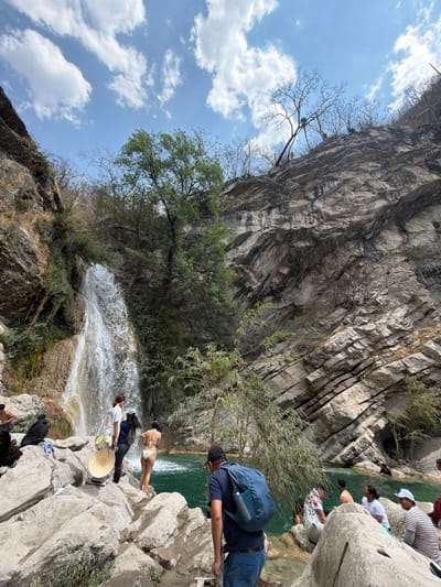 This image depicts a natural recreational area in San Miguel Atlapulco, Mexico, centered around a waterfall and a turquoise pool. The scene is dominated by steep, rocky cliffs on both sides, with the right cliff face exhibiting prominent horizontal stratification. The sky above is clear blue with scattered white cumulus clouds.
On the left, a waterfall cascades down a verdant, rocky cliff into a tranquil, teal-colored pool below. The surrounding vegetation includes both lush green trees and shrubs near the waterfall, and sparser, bare-branched trees on the higher parts of the cliffs, particularly visible on the right slope.
Approximately ten to twelve individuals are present within the scene. In the foreground, positioned to the right, a man wearing a dark blue t-shirt, blue jeans, a black baseball cap, and a blue backpack faces away from the camera, looking towards the water. On the rocky bank near the base of the waterfall, a person with long dark hair, dressed in a white t-shirt and dark pants, stands next to a large, light-colored, circular object resembling a reflector or a drum. Adjacent to them, a woman in a white two-piece swimsuit stands on a rock, facing the water with her back to the camera. Further to the right, several individuals are partially submerged in the turquoise water or seated on the surrounding rocks, engaged in leisure activities. Their attire includes t-shirts, caps, and swimwear. The immediate ground consists of large, light-colored boulders and smaller rocks.